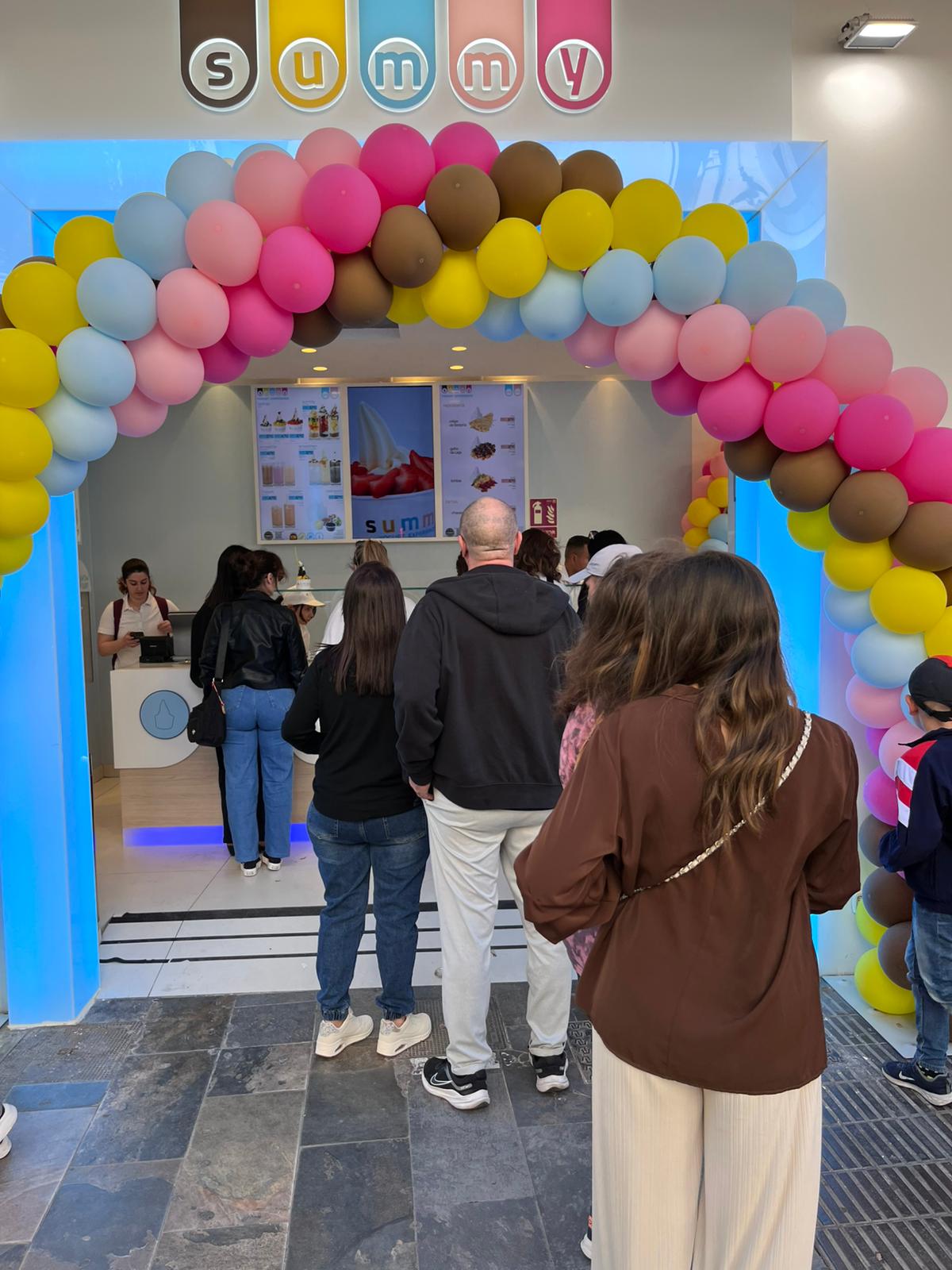 Inauguración de local de yogurt helado Summy con arco de globos de colores y clientes.